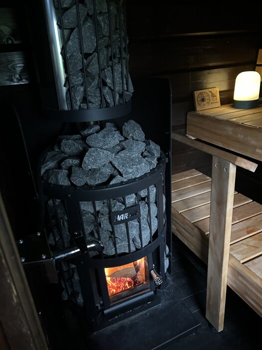 En bastuugn med brinnande ved och stora stenar ovanpå, omgivet av träbänkar.