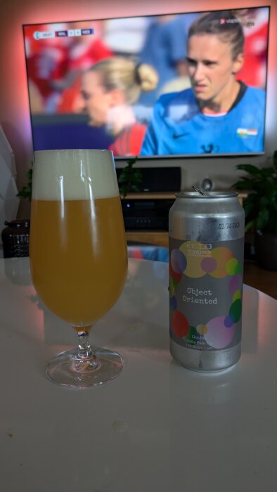 Ölglas och burk med "Object Oriented" öl på bordet framför en TV som visar en fotbollsmatch.