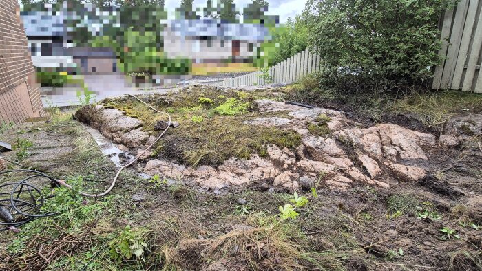 Grävområde med synlig berggrund, gräs och buskar vid sidan om garage eller husfasad, förbereds för bygge eller renovering av betongplatta.