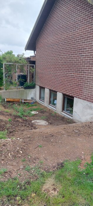 Röd tegelvilla med källarfönster, sluttande mark mot huset, grönska och två synliga brunnslock i marken.