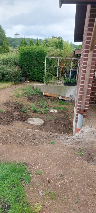 Trädgårdsområde med en sluttning mot en tegelvilla. Två brunnslock syns på marken, omgivna av grönska och en lägre betongmur.