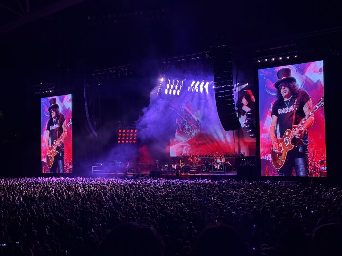 Gitarrist på scen vid en stor konsert, två stora skärmar visar artisten i hatt och solglasögon, publik syns i förgrunden.