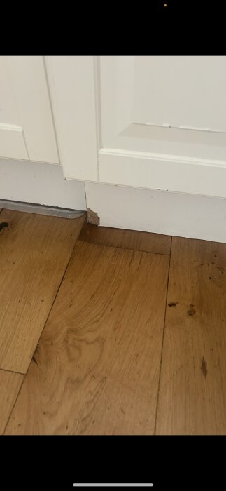 Trägolv med skada vid sockel under en vit kökslucka, cirka 15x15 cm område påverkat.