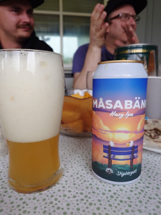Flaska med skummande öl bredvid burk med etiketten "Måsabänk Hazy IPA" från Stigbergets, i bakgrunden två personer i samtal.