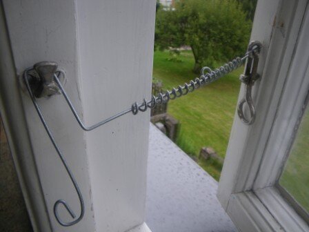 Vädringsbeslag på fönster, spiralformad metalltråd fästad mellan karmen och fönstret, med en gräsmatta i bakgrunden.