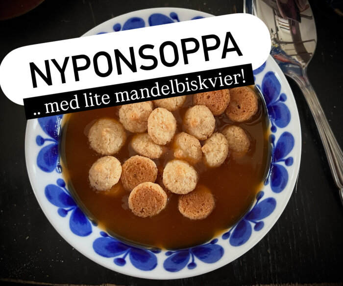 Skål med nyponsoppa toppad med mandelbiskvier, placerad på ett bord bredvid en sked. Vit och blå tallrik.