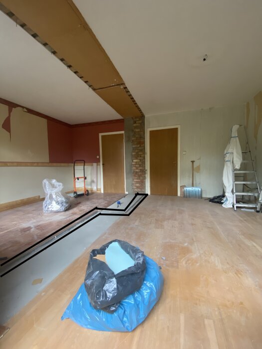 Ett rum under renovering med borttagen garderob, trä- och tegelväggar, stege, plastpåsar på golvet, och en markerad kanal i golvet för framtida arbete.