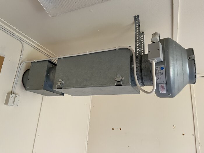 Ventilationssystem monterat i ett rum, med en rektangulär kanal ansluten till en rund fläkt. Bakgrunden visar en vitmålad vägg och tak.