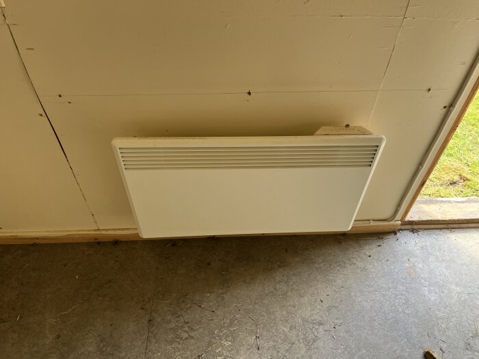 Väggmonterat vitt element vid en vitmålad innervägg, nära ett golv och en öppen dörr med gräs utanför.