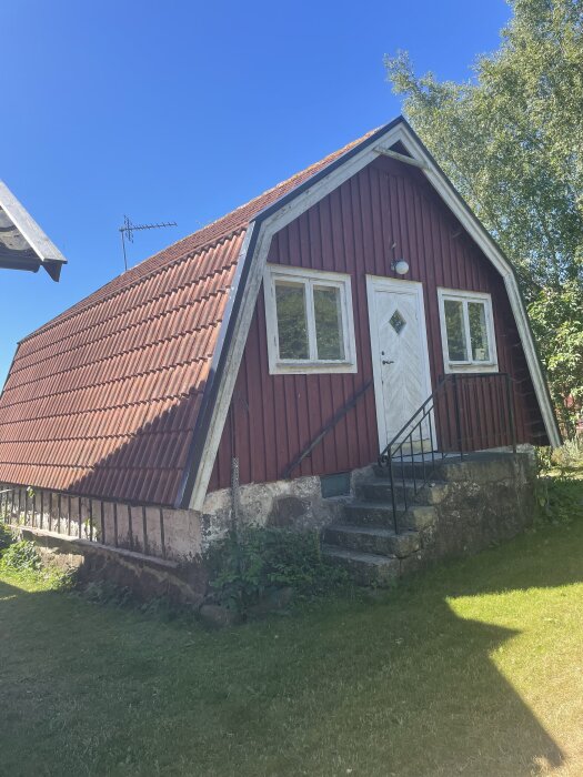 Röd stuga med branta sluttande tak och vita fönster, omgärdad av grönska och belägen på stenfot. Stugan ska vinterbonas och renoveras.