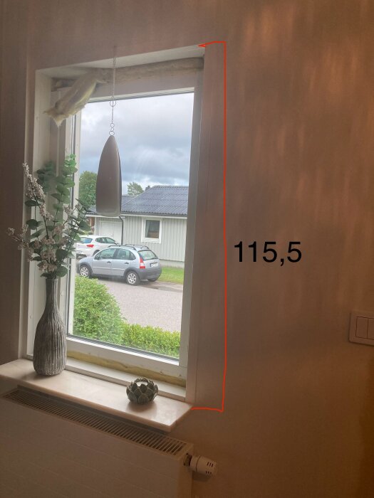Fönster i ett hus med mätning av höjden 115,5 cm markerad med röd linje. Inredning inklusive en vas med blommor och utsikt mot en gata.