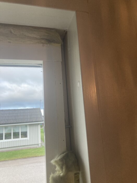 Fönster från insidan med övre bräda borttagen, synlig isolering och skruv, utsikt mot gräsmatta och grå byggnad utanför.