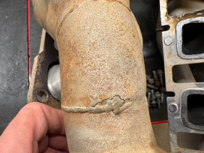 Sprucket avgasrör med rost synligt, hållen av en hand. Bakgrund av svarta och metalliska objekt.