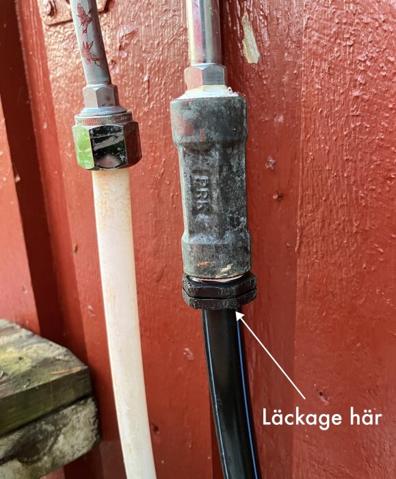 Läckande sommarvattenkoppling med pil som pekar på problemområdet under en mutter, installerad vid en röd trävägg.