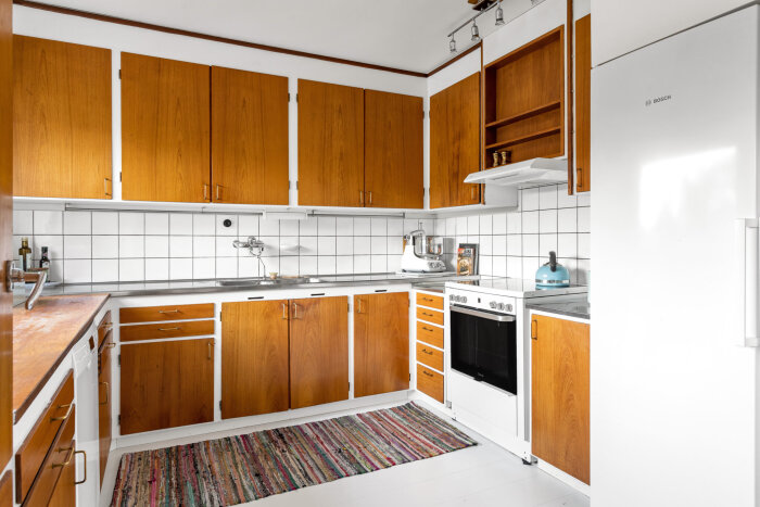 60-tals funkiskök i teak med vita vitvaror, kaklade väggar, synliga överskåp och en färgglad matta på golvet.