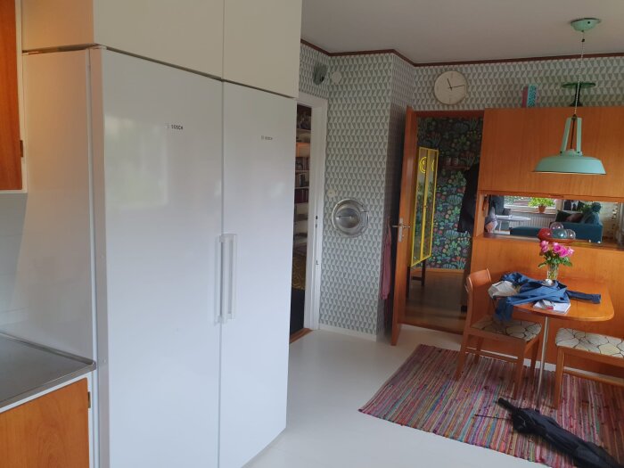 Kök med vita kylskåp, teakinredning och retro tapet. Matbord i teak med stolar, färgglad matta och blommor. Ljusblå lampa i taket.