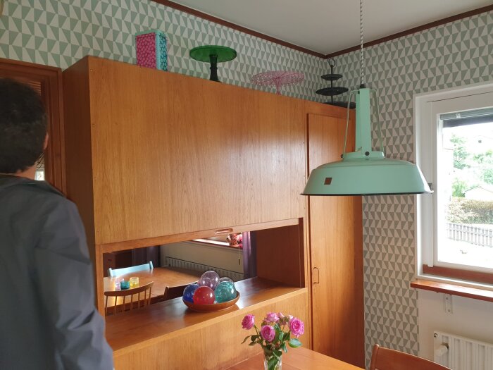 Funkiskök från 60-talet i teak med luckor och ett stort skåp; gröna industrilampa hänger i taket, blommor och färgade glasbollar på bordet.