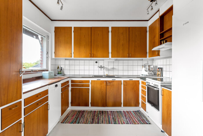 Kök med original teakskåp från 60-talet, vitvaror och vita kakelplattor. En färgglad matta ligger på golvet.