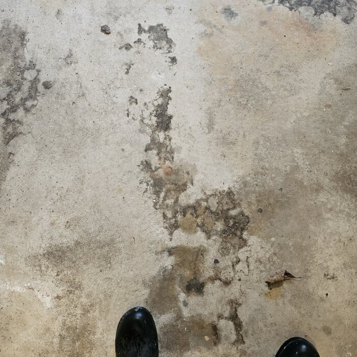 Fläckiga mönster på ett garagegolv av betong, omgivna av damm och en svart sko i bildens nedre kant.