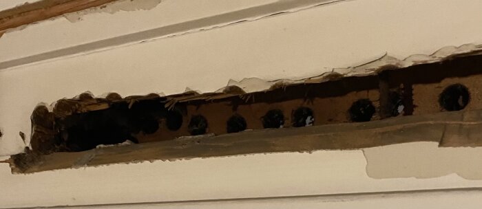 Hål i väggen ovanför fönster med ojämna kanter, synligt trä och små runda ventilationshål.