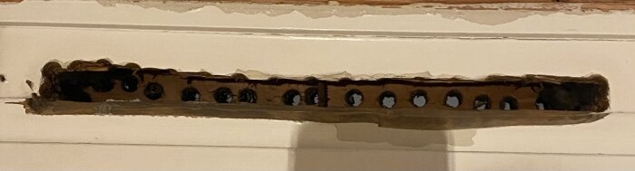 Ett rektangulärt ventilhål ovanför ett fönster, med ojämna kanter och flera små runda hål synliga på insidan.