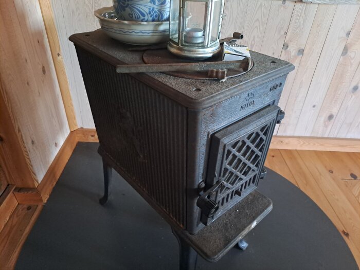 Kamin JØTUL 602 N med stödben till salu, dekorerad med en skål och lykta på toppen; rustik interiörbakgrund.