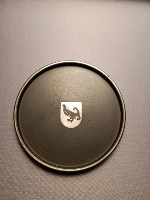 Svart rund skiva av isolit med en silverinläggning föreställande en fågel på en sköld, inspirerad av svensk design och materialutveckling.