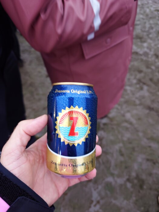 En hand håller en burk Zeunerts Original 3,5% öl med blå och gul etikett på en lerig mark.
