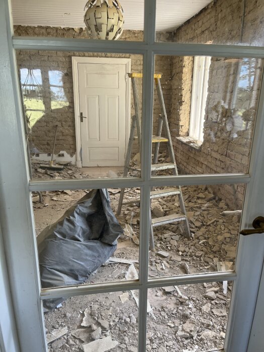 Renoveringsrum med synliga tegelväggar, vit dörr, stege och byggdamm. Plastpåse på golvet omgiven av rivningsmaterial fotograferat genom glasdörr.