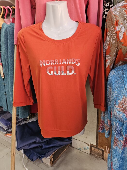 Röd tröja på skyltdocka med texten "Norrlands Guld" i ett klädesstånd.