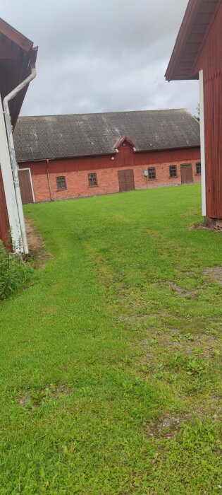 Röd ladugård med tegelfasad omgiven av gröna gräsmattor, en grusväg syns i förgrunden. Molnig himmel i bakgrunden.
