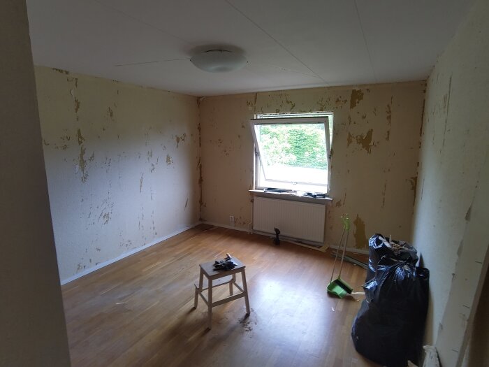 Tomt rum med borttagen tapet, en stol och verktyg på golvet. Fönster öppet och svarta sopsäckar fyllda med skräp bredvid en sopskyffel.