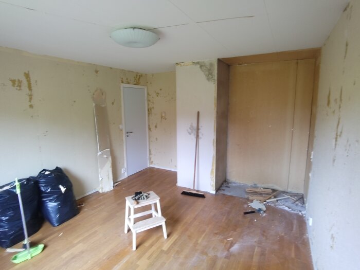 Rum under renovering med borttagen tapet, trästegpall, sopsäckar och redskap. Spår av tidigare installation syntes där vägg reviderats.