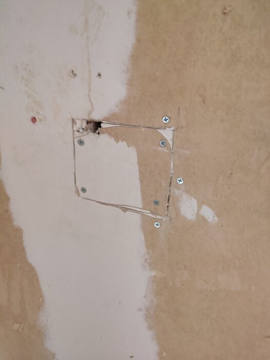 Vägg med påbörjad renovering, synliga skruvar och rektangulärt hål avlägsnat för värmepumpsinstallation.