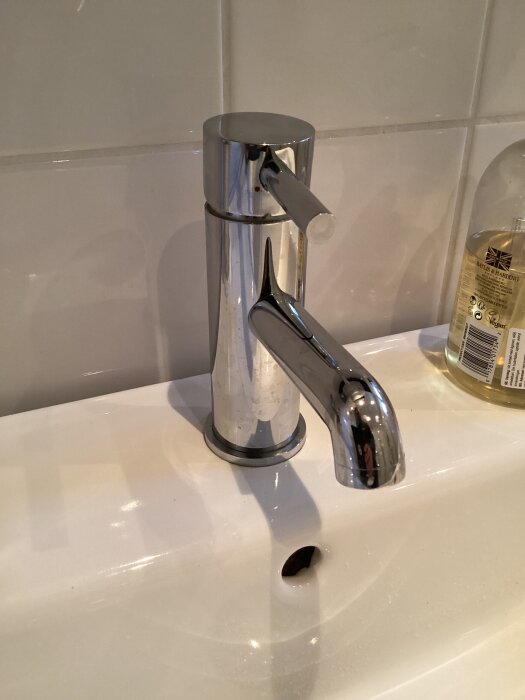 Kromad enhandstermostatblandare med runt handtag och pip på vit tvättställ, badrumsvägg i kakel och en flaska handtvål.