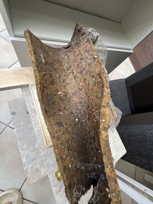Isolermaterial med fukt- och mögelfläckar, delvis täckt av plast, placerat i en inomhusmiljö med kakelgolv och möbler i bakgrunden.