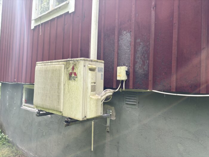 Fasad av faluröd Asfaboard med luftvärmepump monterad på väggen; synliga fuktskador från vattenstänk under pumpen.