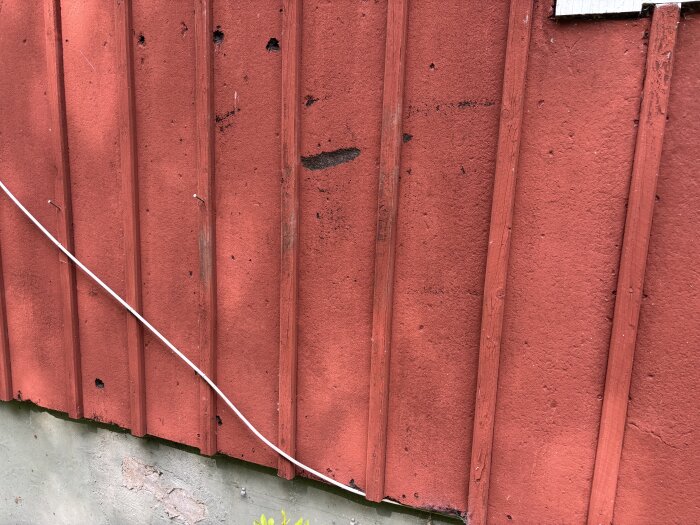Röd fasad i Asfaboard med synliga skador och mörka fläckar nära grunden; en vit kabel löper diagonalt över panelen.