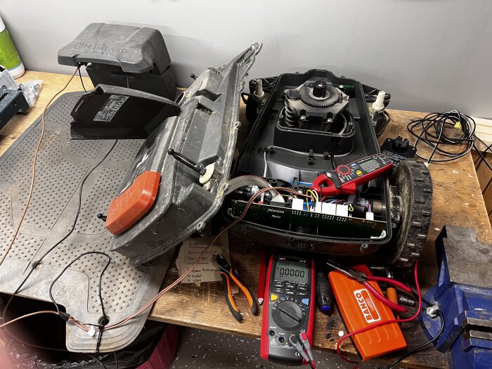 Öppen robotgräsklippare på arbetsbänk med kretskort exponerat, omgiven av multimeter och verktyg som tänger och skruvmejslar.