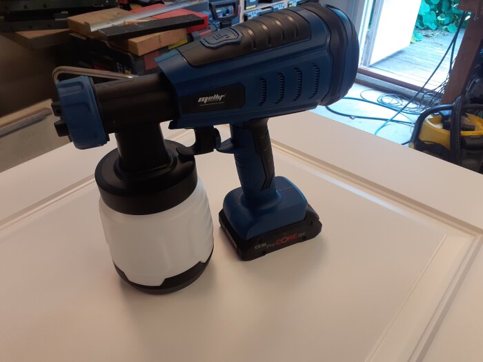 Blå färgspruta på vit dörr i garage, avsedd för Boschbatterier, med tre munstycken och justerbara inställningar för sprutmönster och mängd.