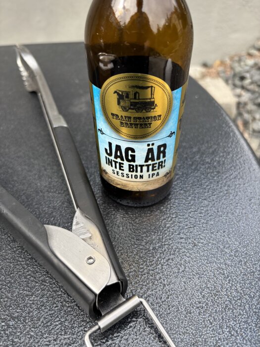 Flaska med etikett "Jag är inte bitter" Session IPA bredvid en grilltång på ett svart bord.