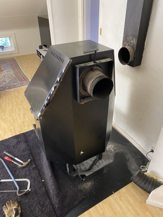 Svart plåtkamin står på svart matta i ett rum med trägolv; verktyg, som en bågfil och hammare, ligger framför kaminen.