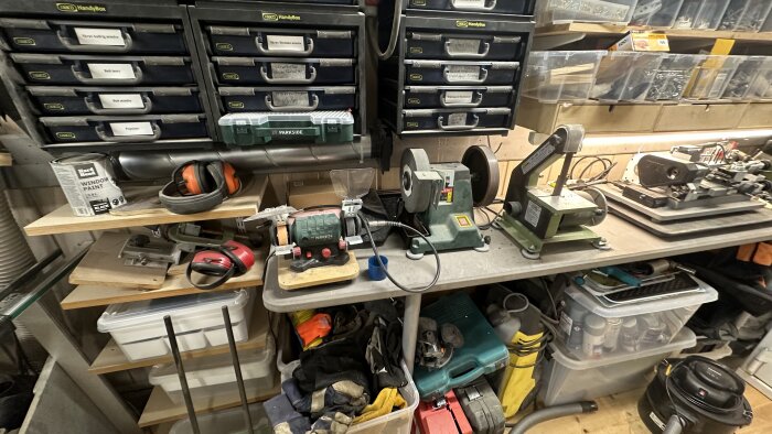 Verkstadshörna med verktyg som slipmaskiner, hyllor med förvaringslådor, färgburk, hörselskydd och diverse material och utrustning.