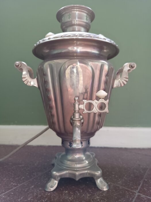 Elektrisk samovar märkt CCCP från 1965, stående mot en grön vägg med två handtag och en kran.