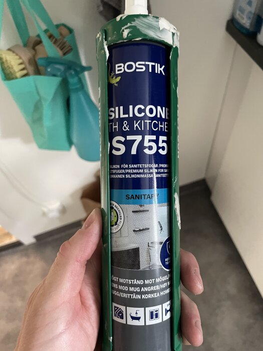 En hand håller i en tub Bostik S755 silikon för bad och kök med etikett i blått och grönt, avsedd för sanitetsfogning.
