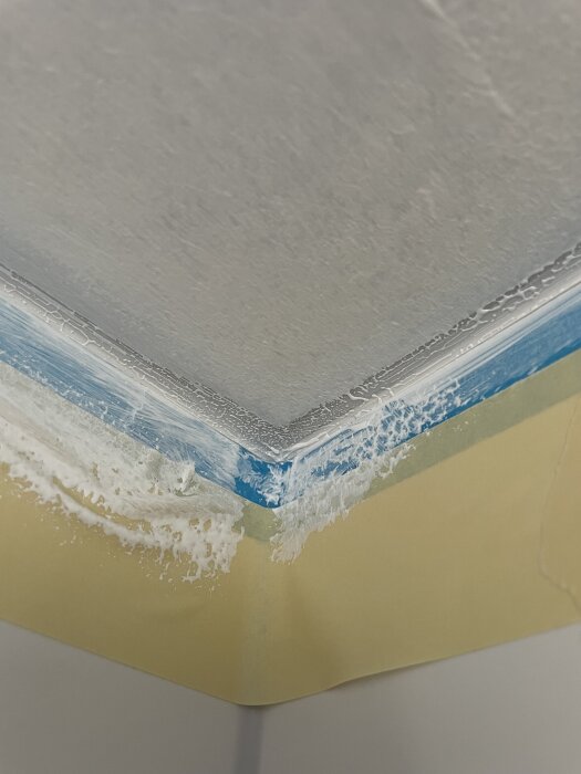 Hörn av badrumsvägg med silikonfog som är svår att måla över, med synlig blå maskeringstejp och vit färg som inte fäster bra.