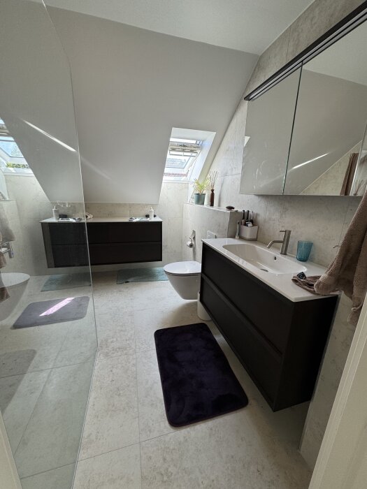 Modernt badrum med snedtak, granitkeramiska plattor på golv och väggar, svart kommod och spegelskåp, väggmonterad toalett och två takfönster.