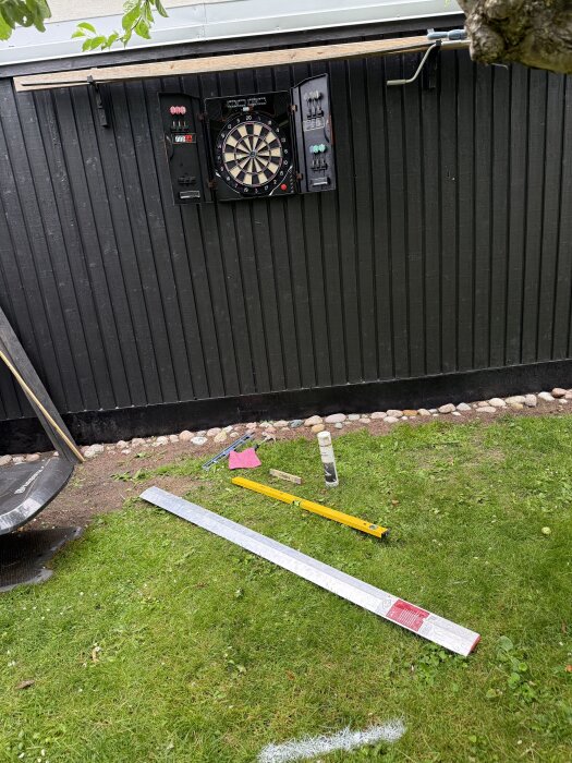 Utomhus darttavla monterad på en svart vägg med dartpilar. Verktyg inklusive ett vattenpass ligger på gräsmattan nedanför.