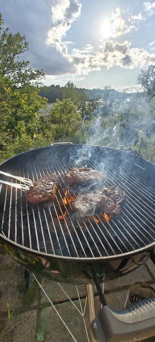Grill som brinner, stekar ligger på gallret med rök stiger upp och en klar himmel i bakgrunden.