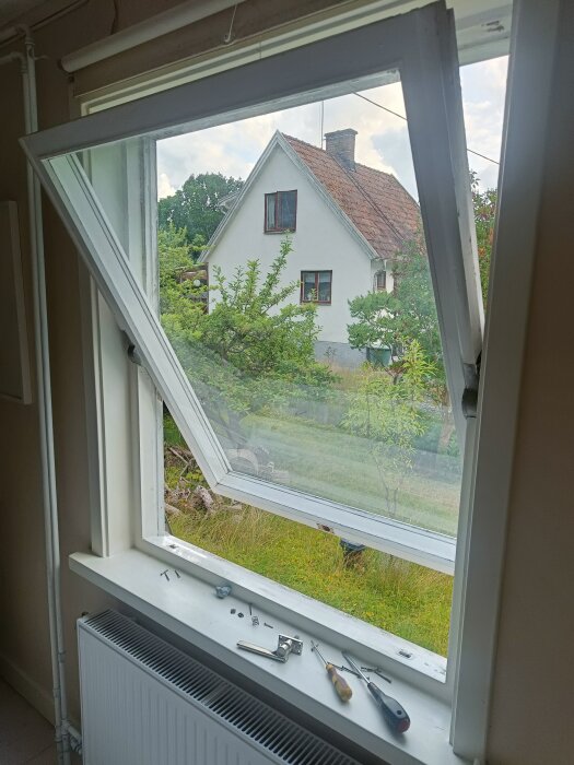 Öppet fönster med verktyg och trasiga delar på fönsterbrädan, utsikt mot trädgård och hus i bakgrunden.